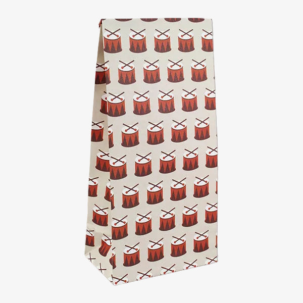 A tall, narrow paper bag or gift bag with a repeating pattern of red and black drums and drumsticks on a light beige background, shown against a plain white studio backdrop.