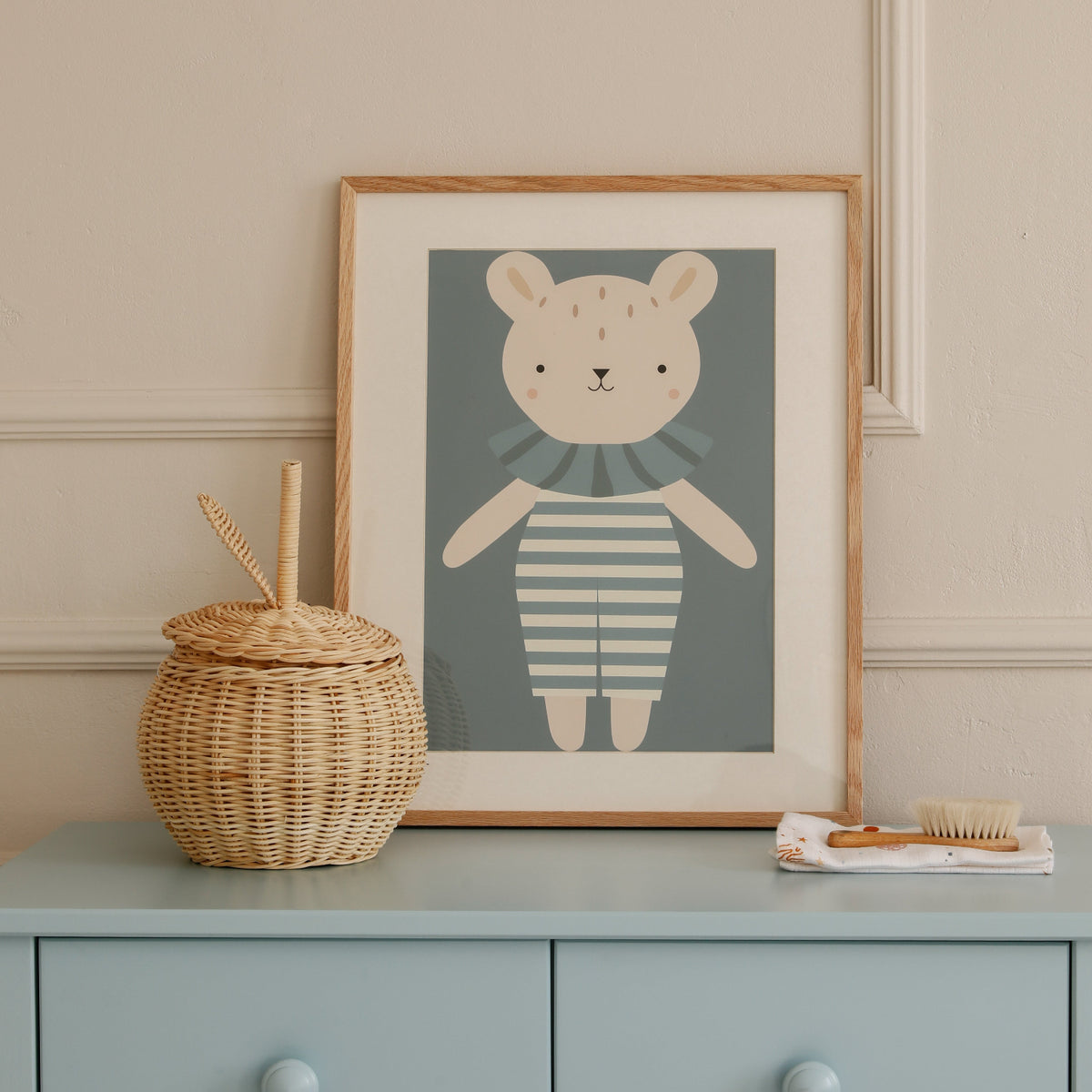 A framed art print of a cute cartoon bear wearing a striped shirt, styled on a dusty blue dresser next to a woven basket and small accessories.