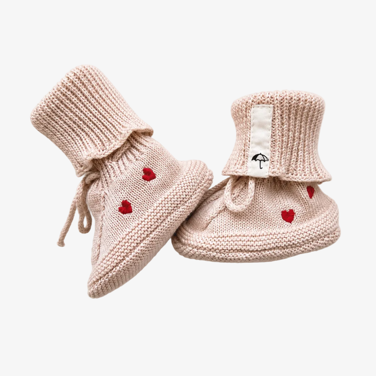 A pair of beige knit baby booties with small red heart details and a white brand tag featuring a black umbrella logo, displayed on a light cream background.