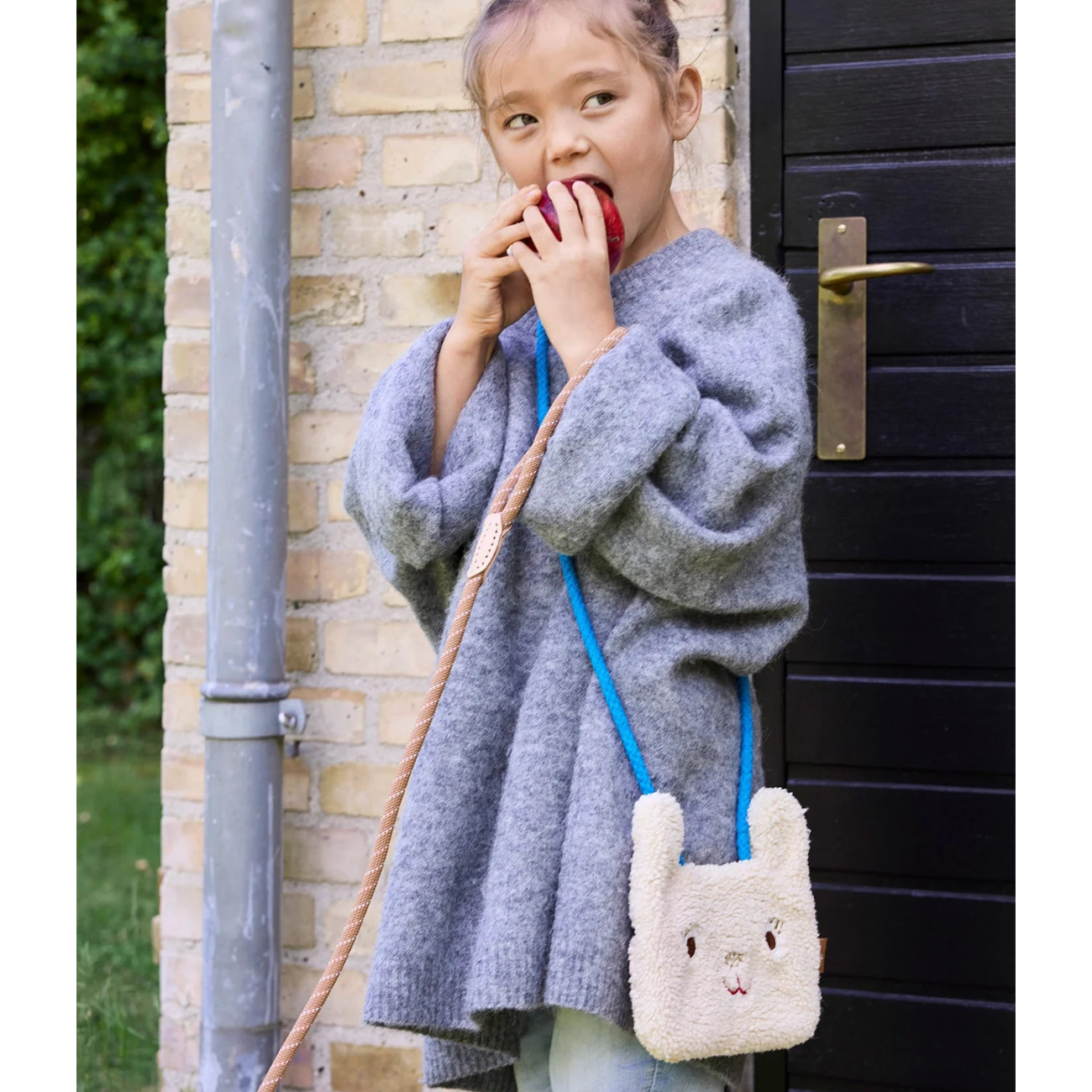Child wearing a gray sweater with a fluffy bunny bag, standing against a brick wall.