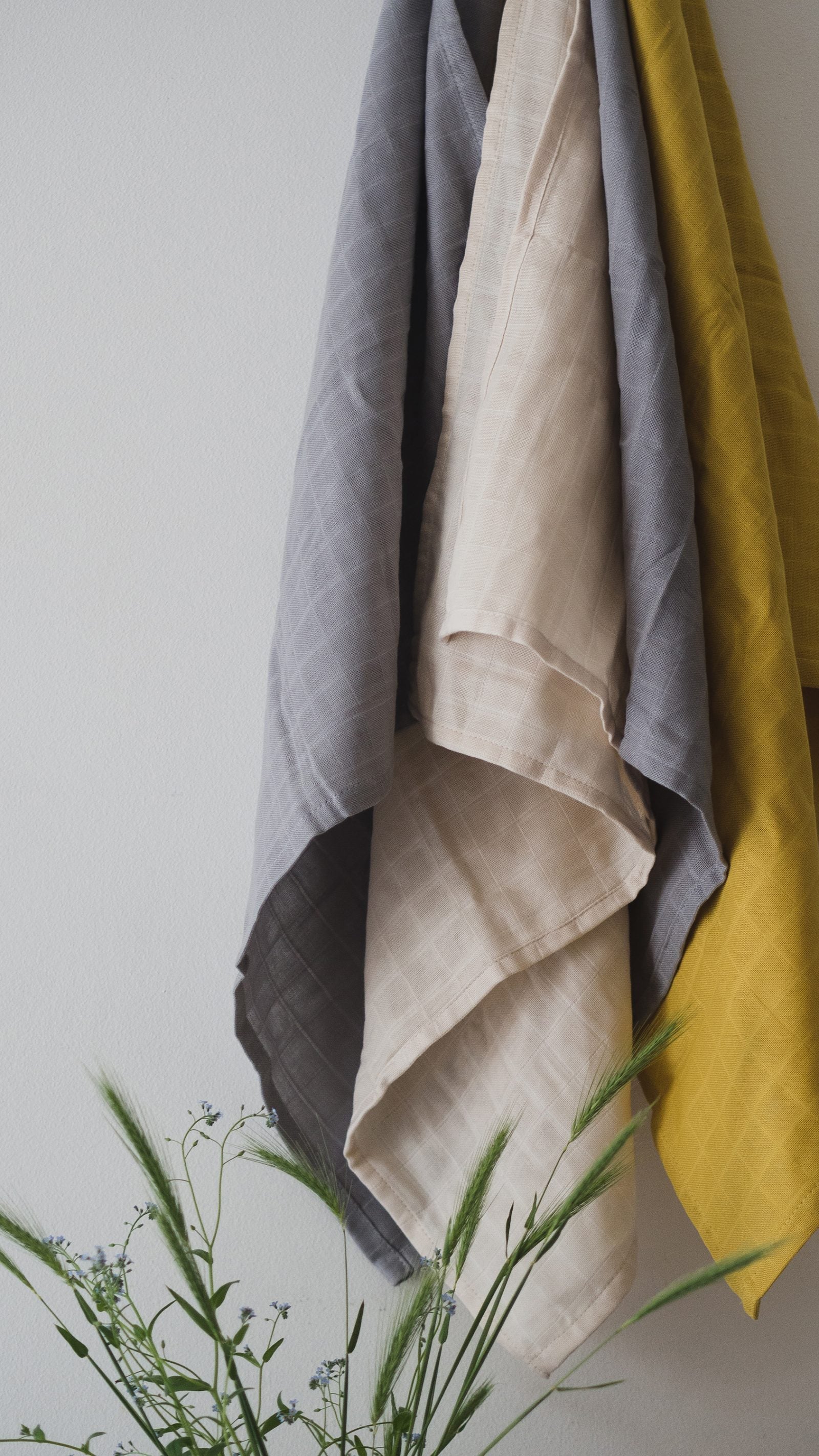 Three towels in gray, beige, and yellow hanging against a white wall with green plants at the bottom.