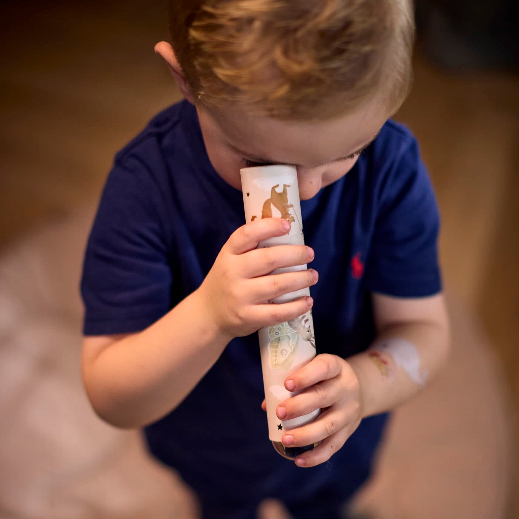 Child holding a cylindrical object with a pattern, standing on a wooden floor. 