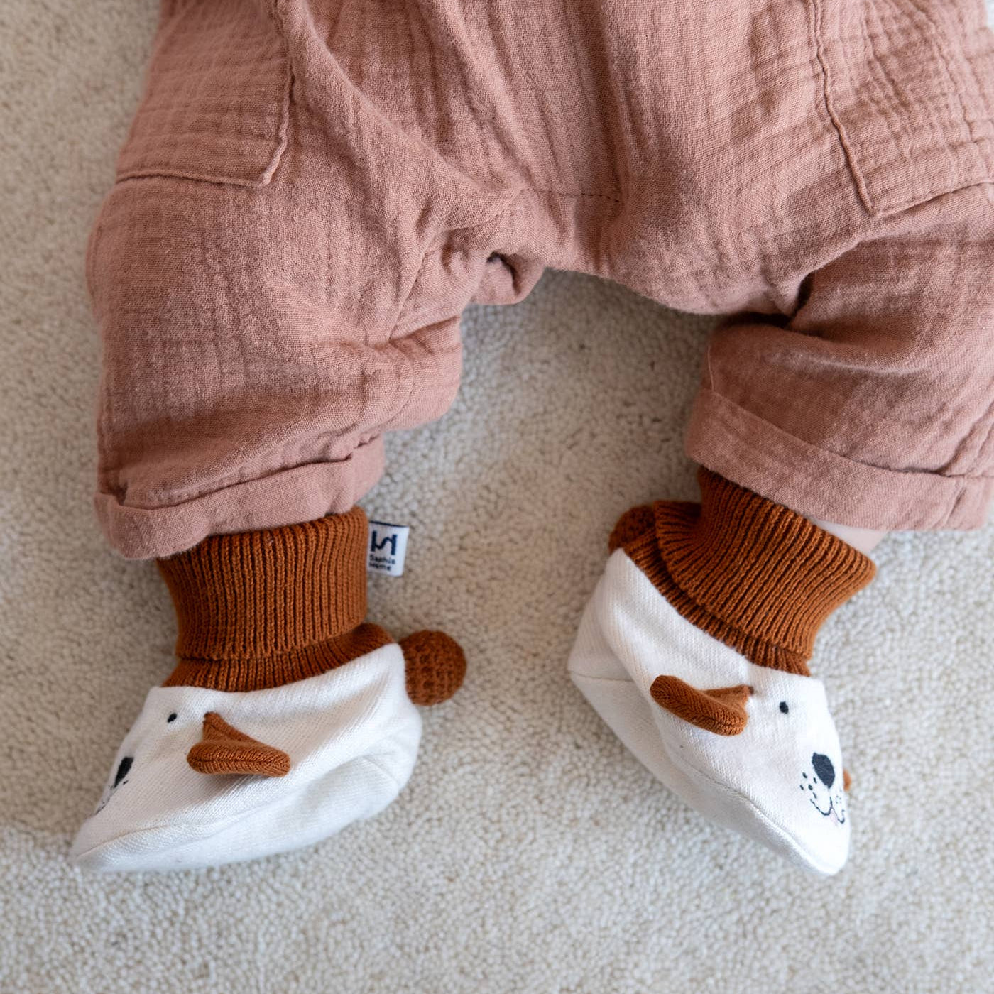 A close-up of a baby's legs wearing dusty rose muslin overalls, rust brown socks, and white bootie-style socks with brown ear details, laid on a light beige carpet.