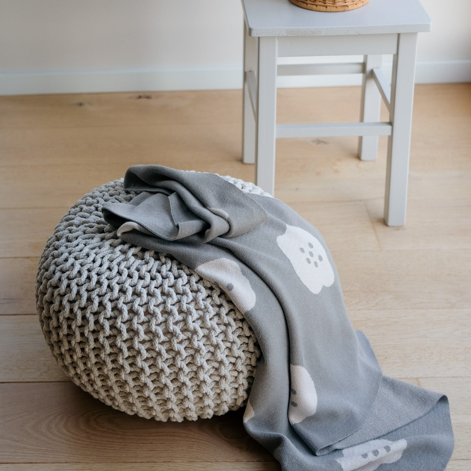 Knitted pouf with a gray blanket featuring cloud patterns on a wooden floor.