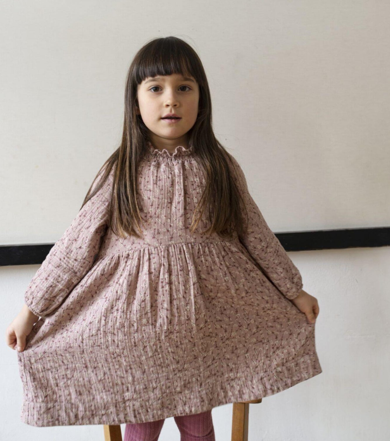 Young girl wearing a pink floral dress sitting on a stool against a plain background