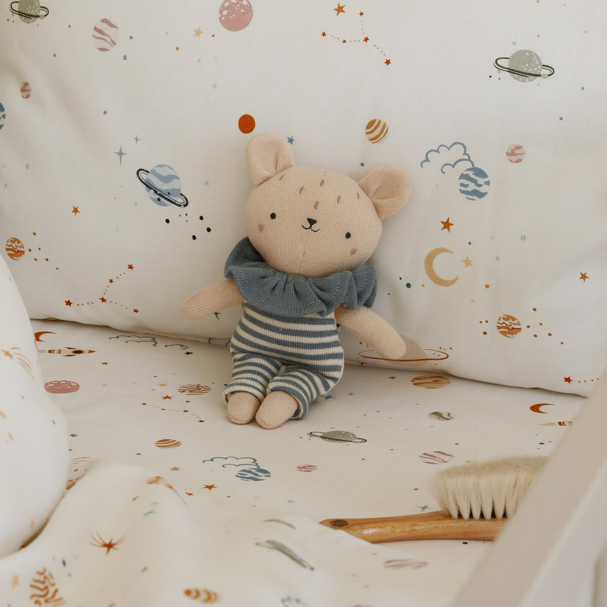 A cozy baby crib setup featuring a white bedding set with a colorful space-themed print of planets, stars, and moons, styled with a plush teddy bear and a wooden baby brush.