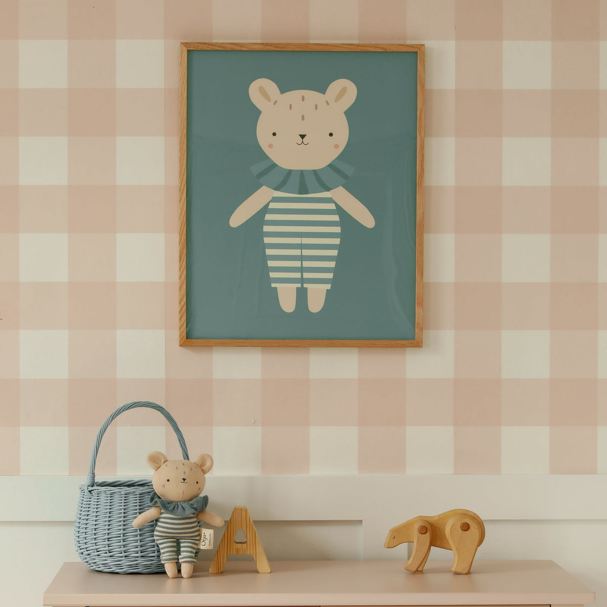 A framed art print of a teddy bear in a striped outfit hanging on a pink and white checkered wall above a light pink wooden dresser with toys on top.
