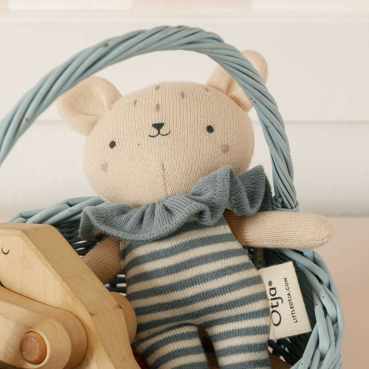 A beige knitted teddy bear doll wearing a blue-gray striped outfit and ruffled collar, sitting inside a light blue wicker basket with a wooden toy partially visible.