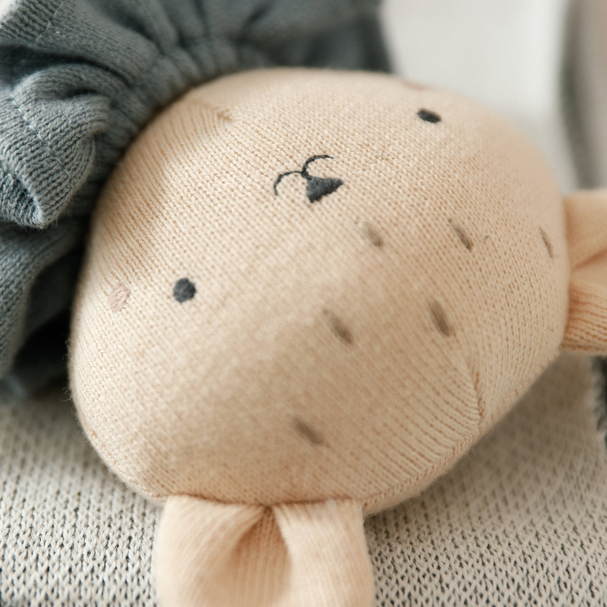 A close-up of a soft knitted plush toy with a round beige face, small embroidered black facial features, a folded cream ear, and a dark gray fabric hair accessory resting on top.