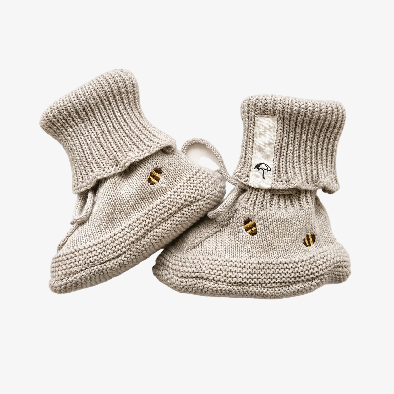 A pair of light beige knit baby booties with small embroidered bee details and a white fabric cuff tab featuring a black umbrella logo, set against a plain off-white background.