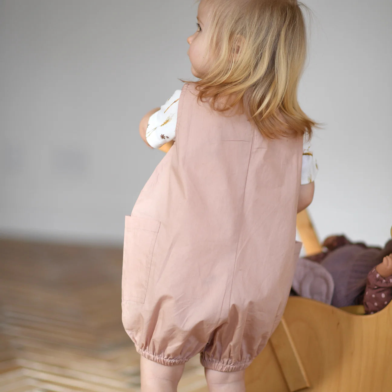 Child wearing a pink romper standing on a wooden floor.